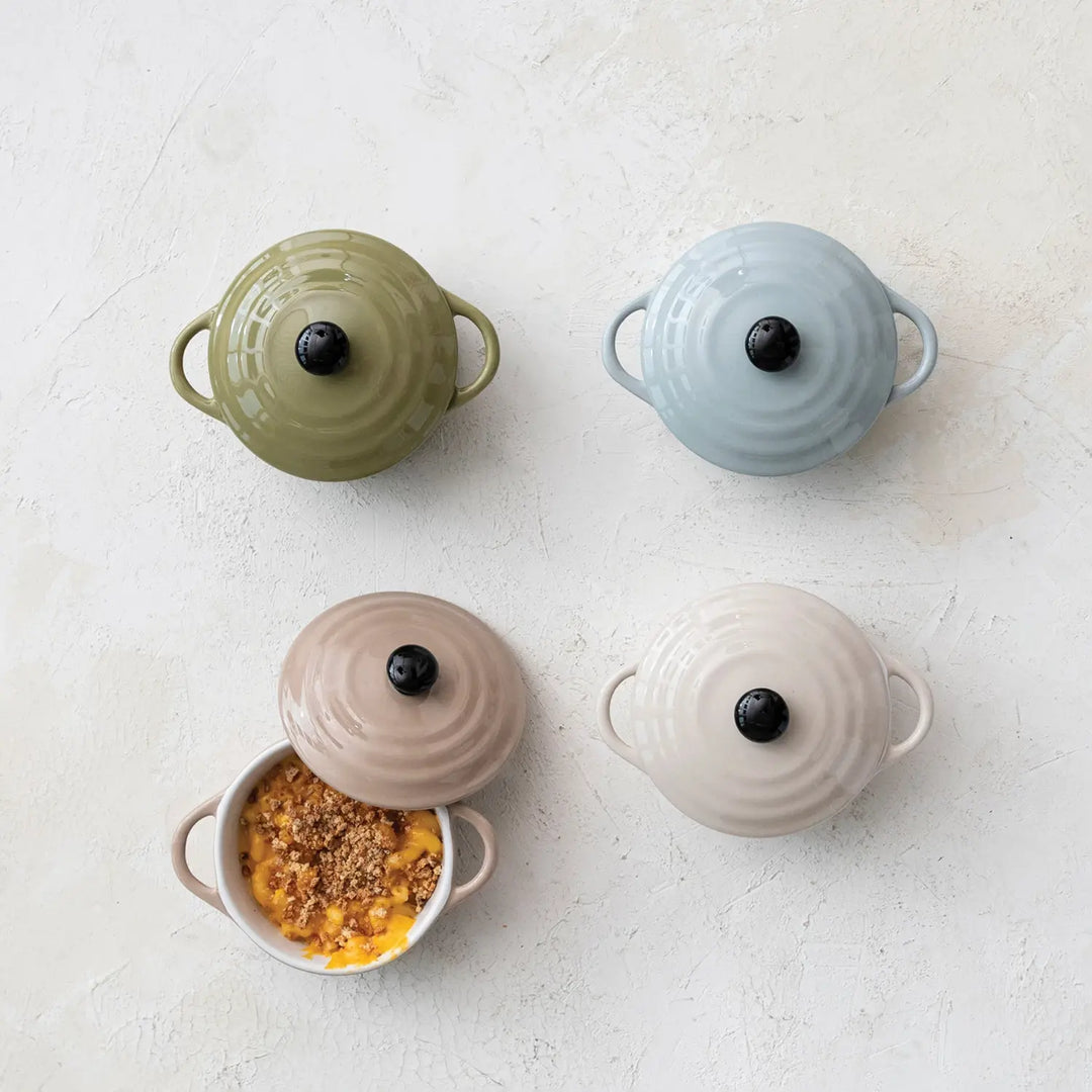 Four 1-cup stoneware mini bakers with lids in olive, blue, tan, and cream, one filled with food.