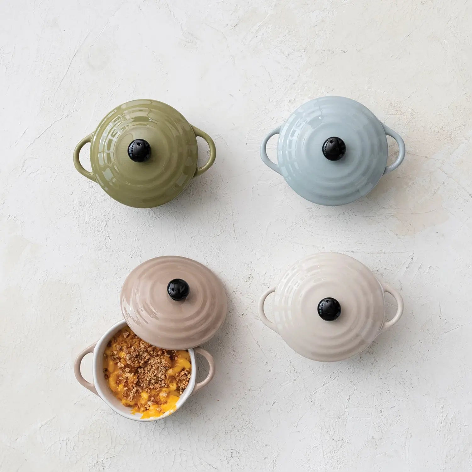 Four 1-cup stoneware mini bakers with lids in olive, blue, tan, and cream, one filled with food.