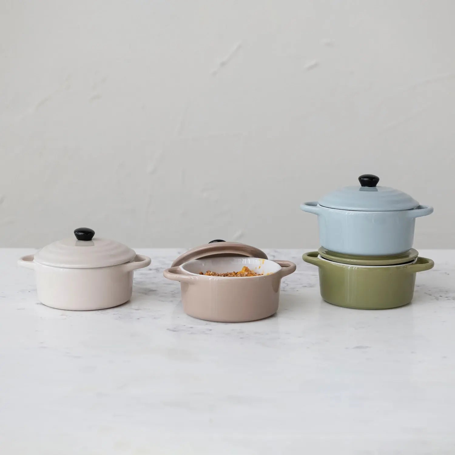 Four colorful stoneware mini bakers with lids arranged on a white marble surface.