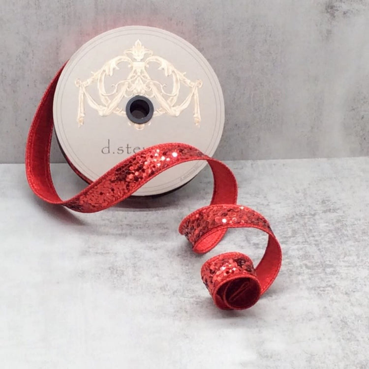 A spool of 1-inch wide red glitter ribbon with a sequined texture, curled on a grey surface.