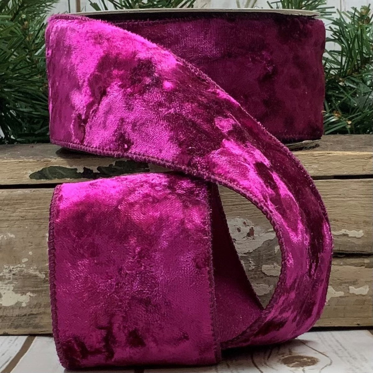 2.5 inch wide fuchsia crushed velvet ribbon on a wooden surface with pine branches.