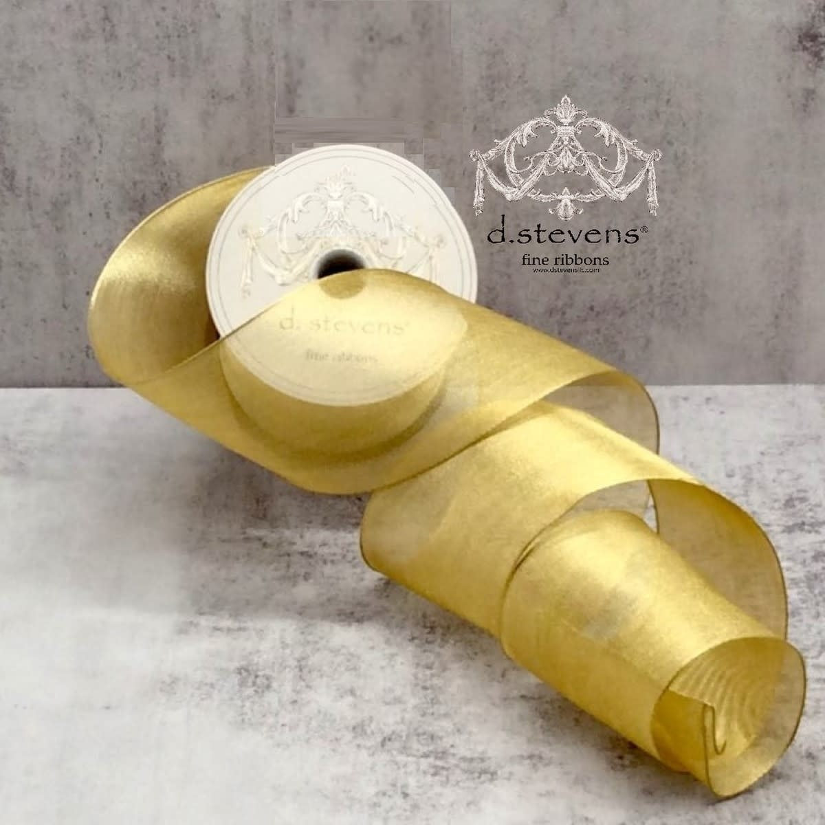 A roll of gold metallic mesh ribbon by d.stevens on a grey textured surface.