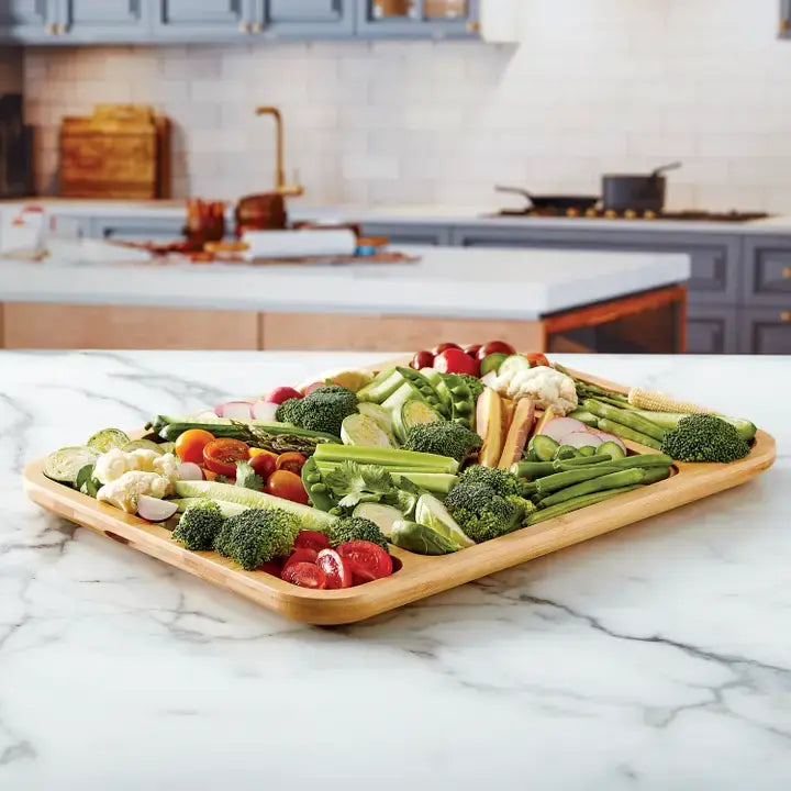 Bamboo cutting board with three compartments filled with fresh vegetables on a marble countertop.