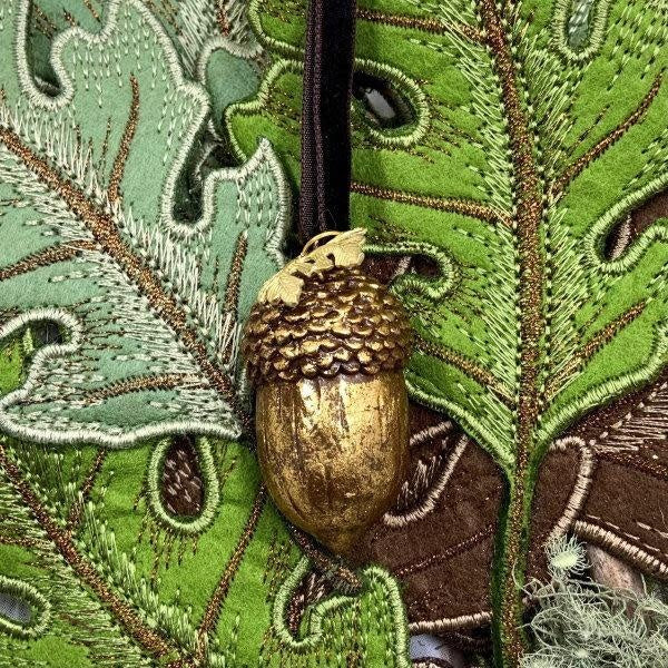 3-inch antique gold resin acorn ornament hanging against a background of embroidered green leaves.