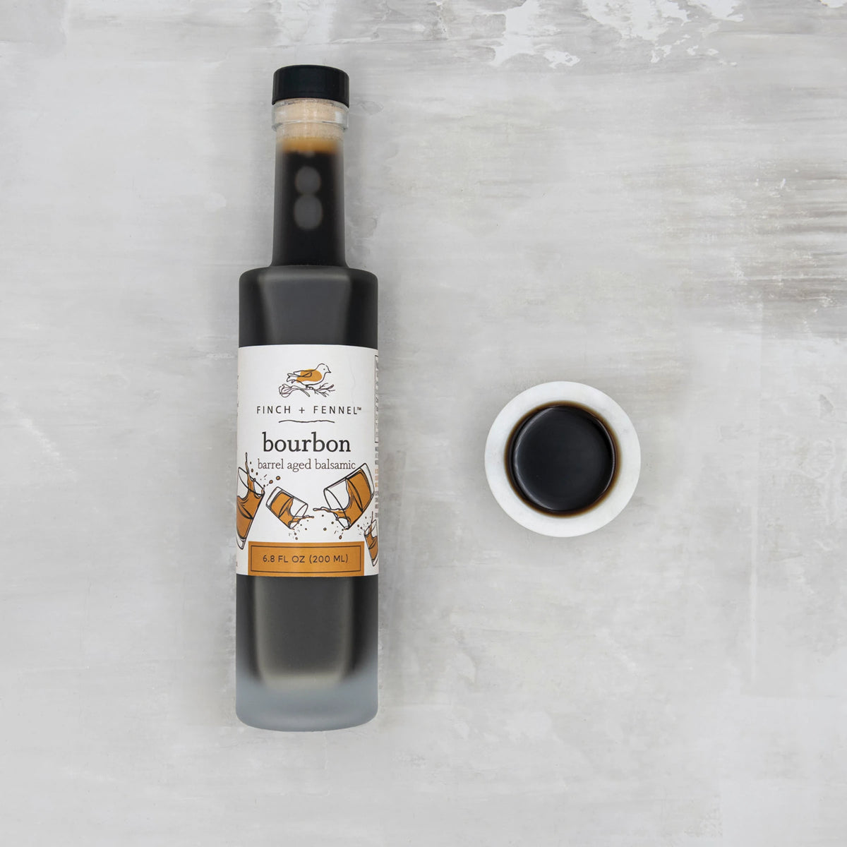 A 6.8 oz bottle of Finch + Fennel Bourbon barrel aged balsamic next to a small bowl of vinegar.