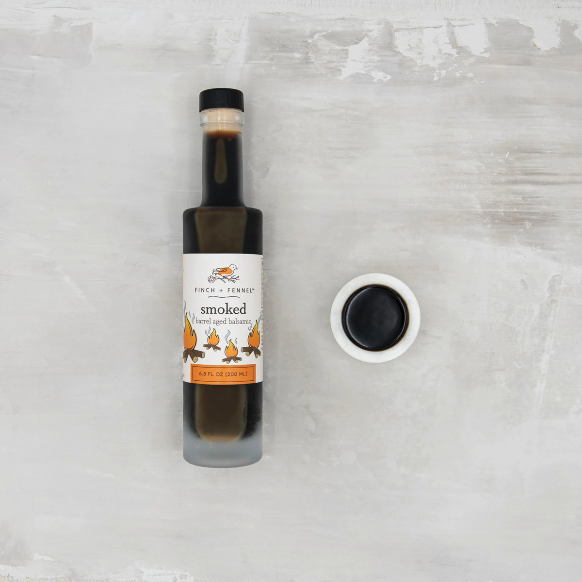 A bottle of Finch + Fennel Smoked Barrel Aged Balsamic next to a small bowl of balsamic vinegar.