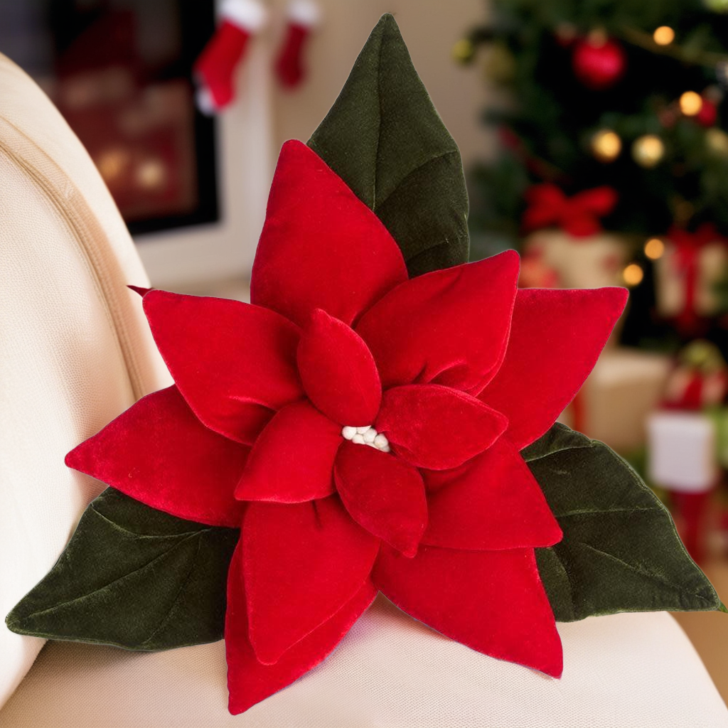 Red velvet poinsettia flower pillow with green leaves, resting on a cream-colored surface.
