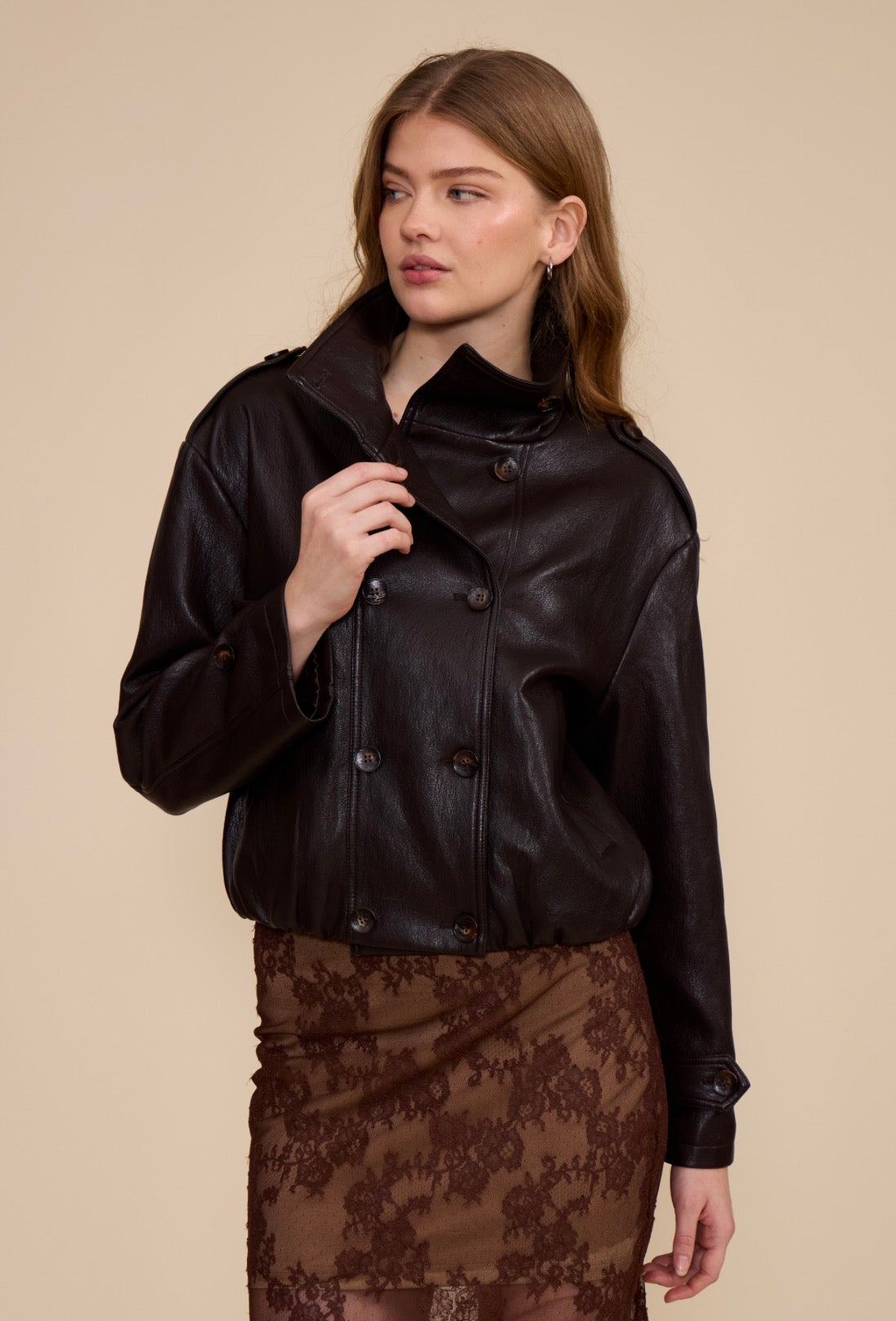 A woman wearing the Corina Funnel Neck Jacket in dark brown faux leather with a lace skirt.