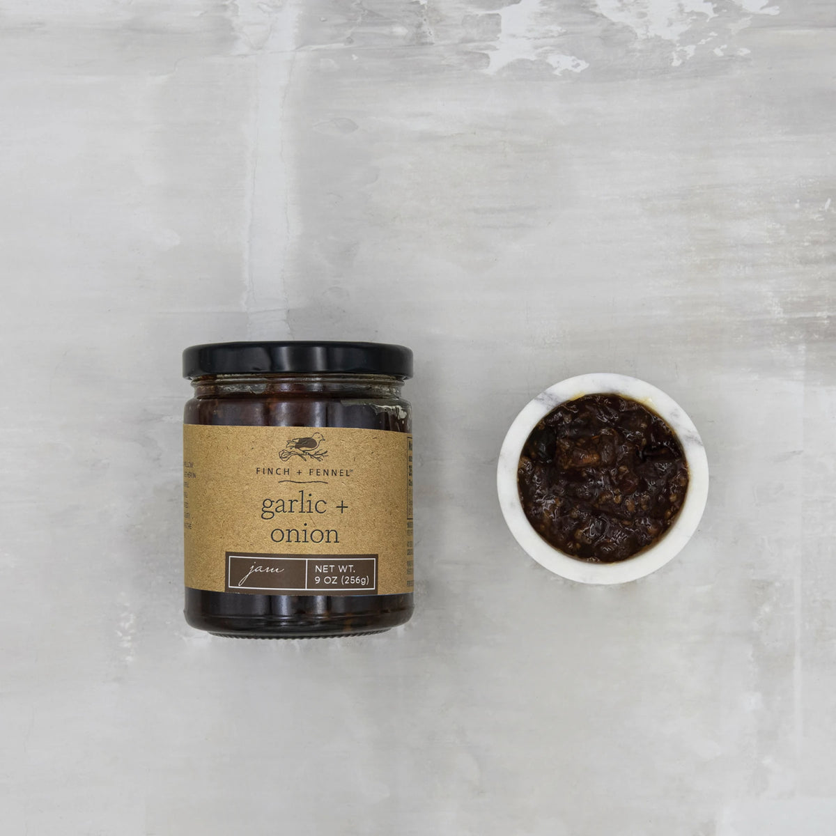 A 9 oz jar of Finch + Fennel Garlic + Onion Jam next to a small white bowl of the dark savory jam.