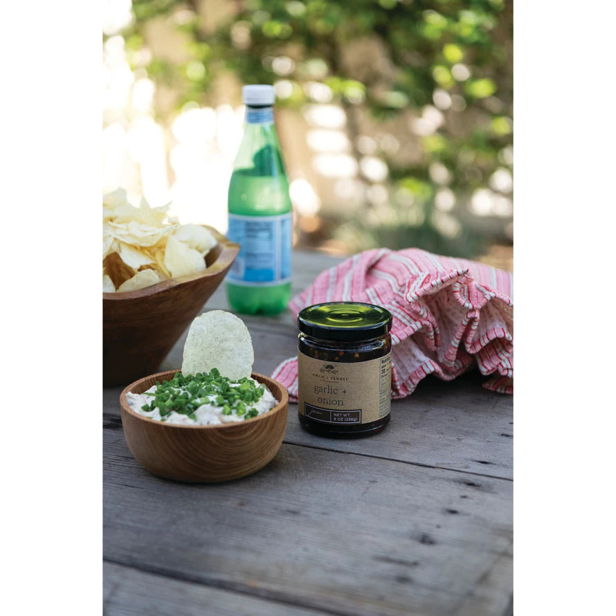 A jar of Garlic + Onion Jam on a wooden table next to a bowl of dip topped with chives and chips.