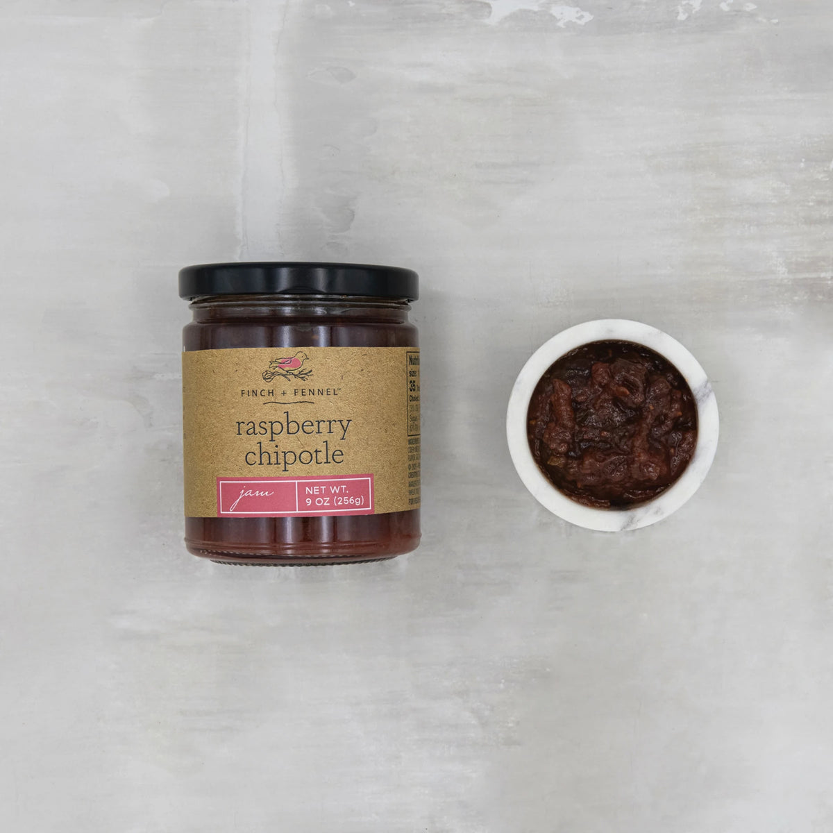 A 9 oz jar of Finch + Fennel Raspberry Chipotle Jam next to a small white bowl of the jam.