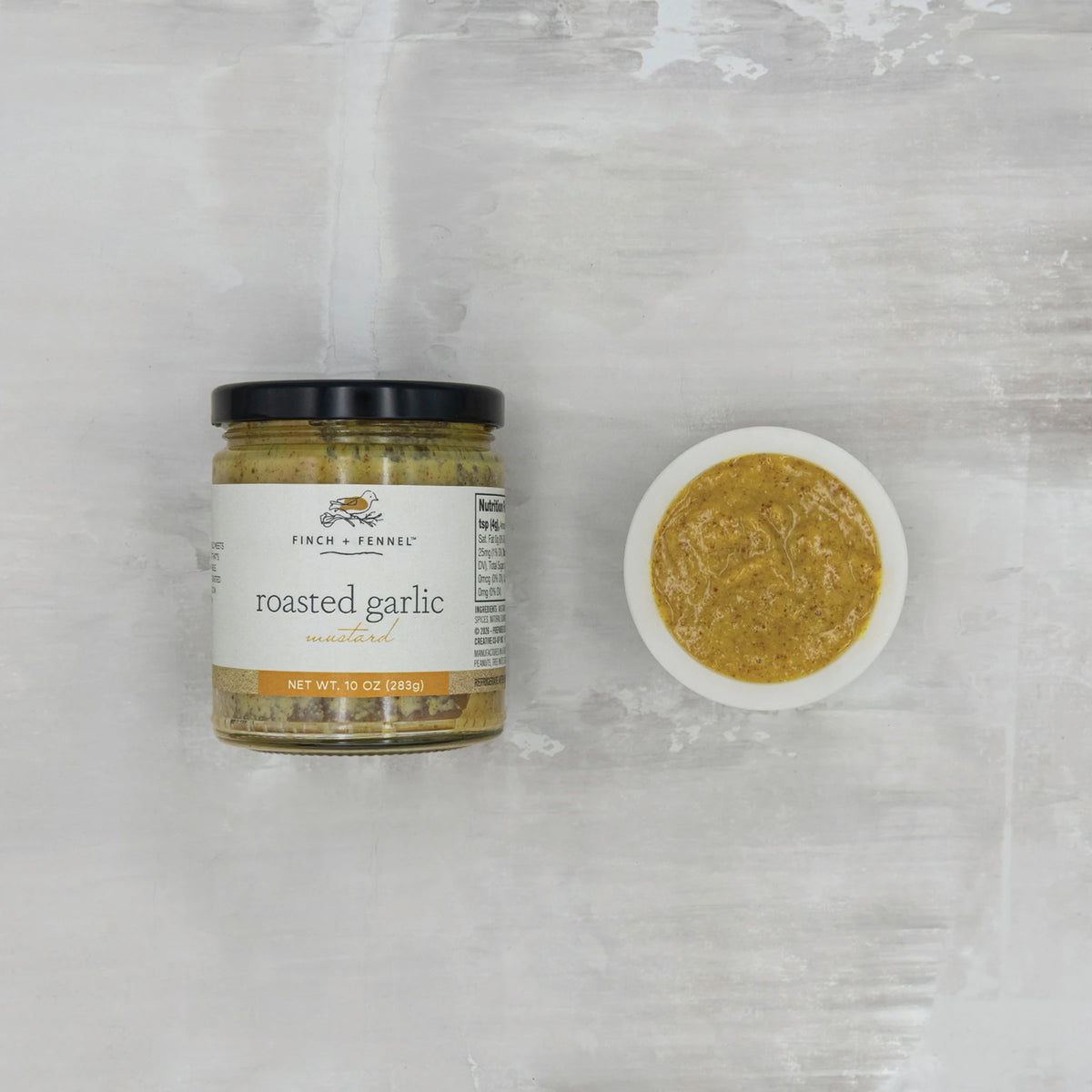 A jar of Finch + Fennel Roasted Garlic Mustard next to a small white bowl of the mustard.