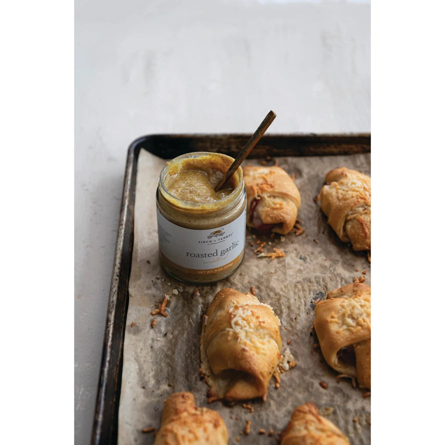 9 oz. Roasted Garlic Mustard in a glass jar on a baking sheet with golden crescent rolls.