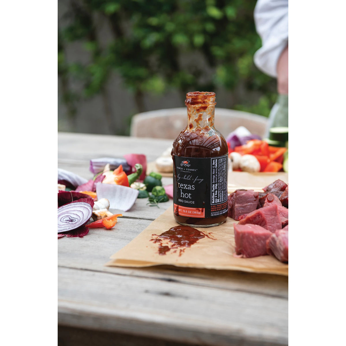 A bottle of Texas Hot BBQ Sauce on a wooden table with raw meat, onions, and peppers.