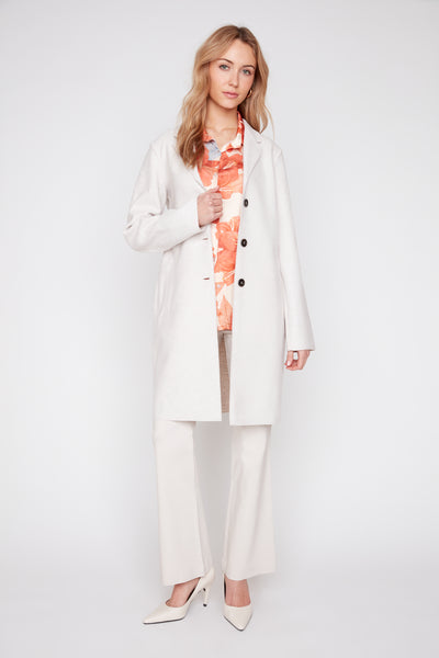 Woman wearing a long oatmeal brushed knit coat with raw edges over a floral shirt and white pants.