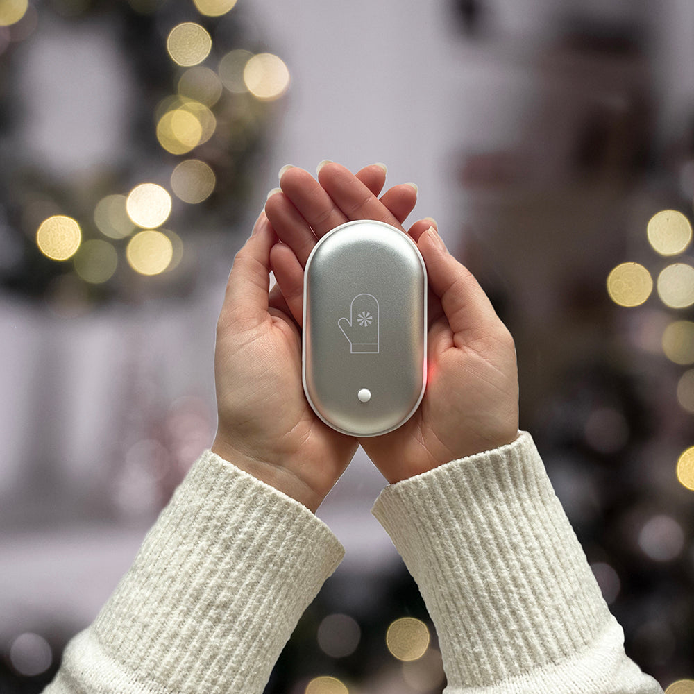 Silver oval hand warmer with a mitten icon and a small indicator light, held between two hands.