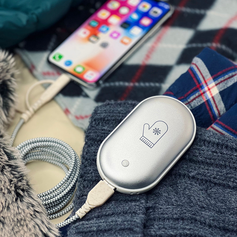 Silver oval hand warmer with a mitten icon, charging via USB cable, resting on a dark gray knit sweater.