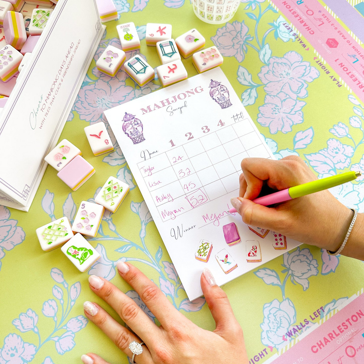 A person writing on a Mahjong Scorecard Pad with Taylor Tiles and a pink and green pen.