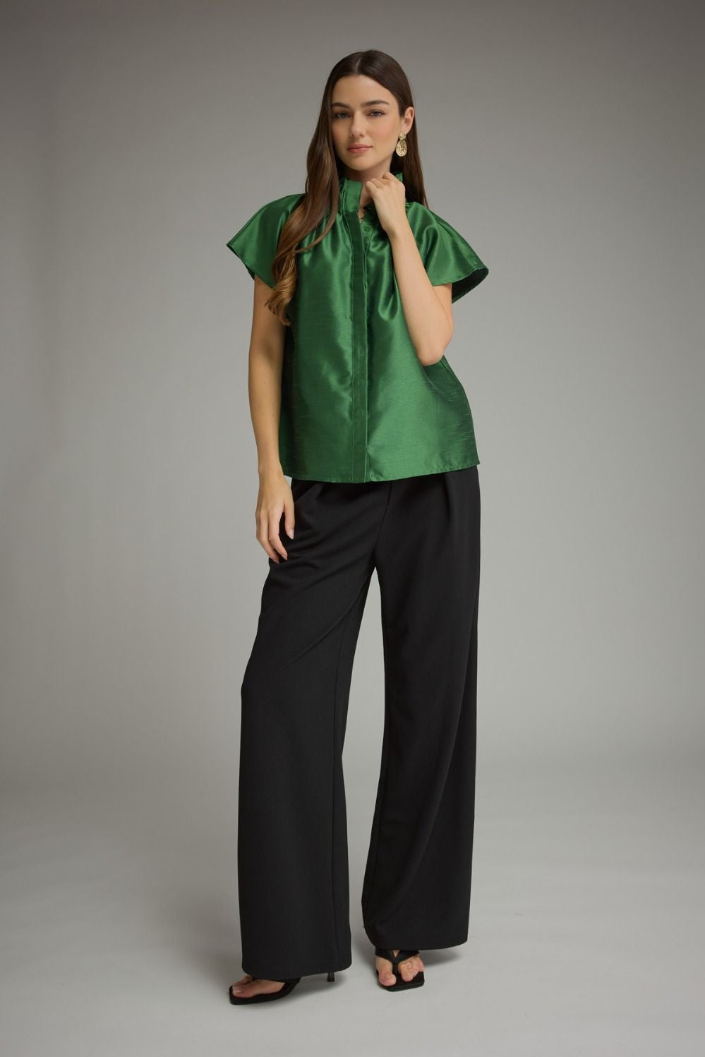 Woman wearing a green Maya solid cap sleeve top with a ruffled high neckline and black trousers.