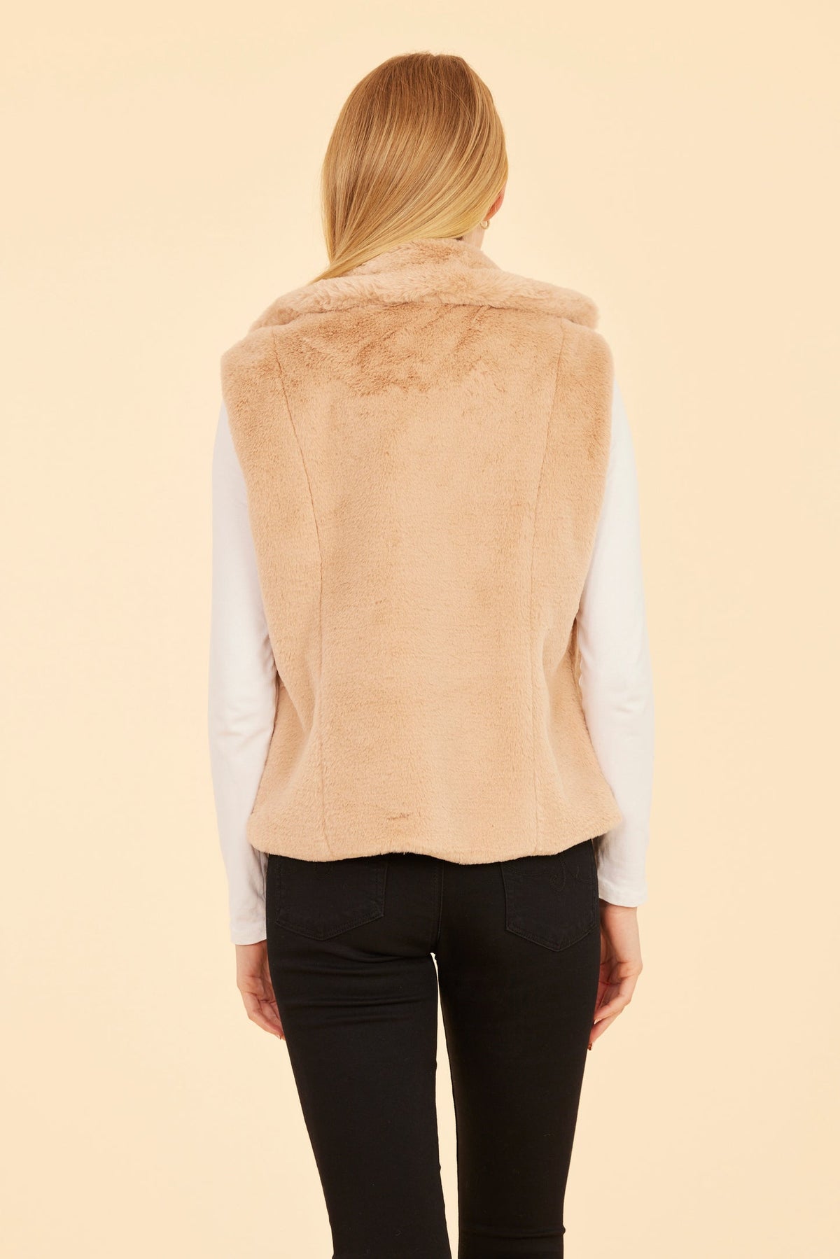 Back view of a woman wearing a beige plush faux fur vest with a notched collar and black pants.