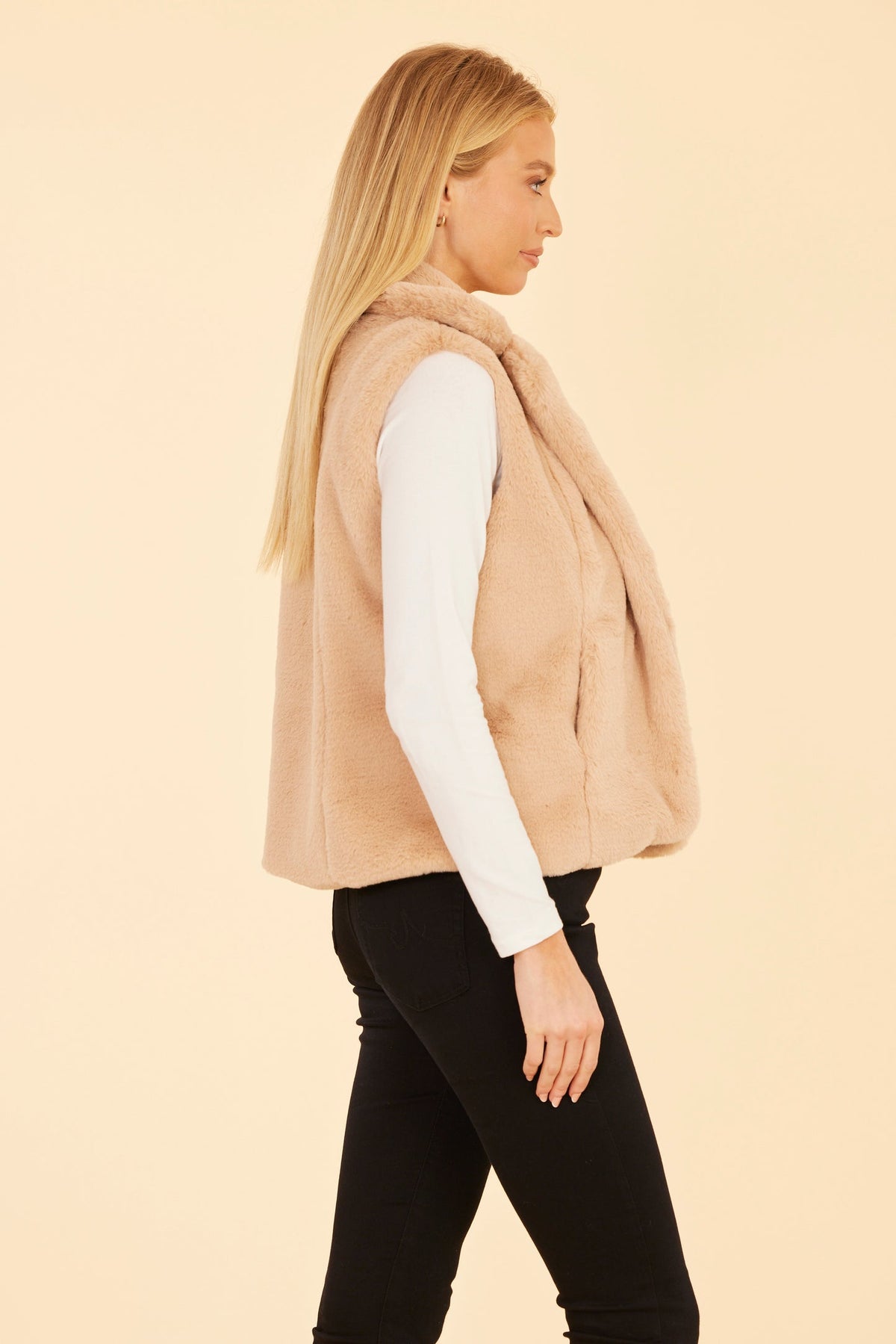 Side view of a woman wearing the Naomi plush faux fur notch collar vest in a light tan color.