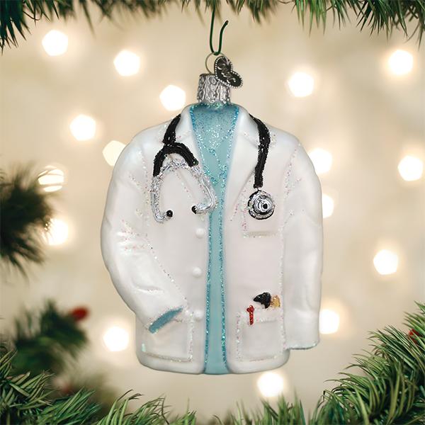 Glass doctor's coat Christmas ornament with a stethoscope and blue scrubs hanging on a tree.