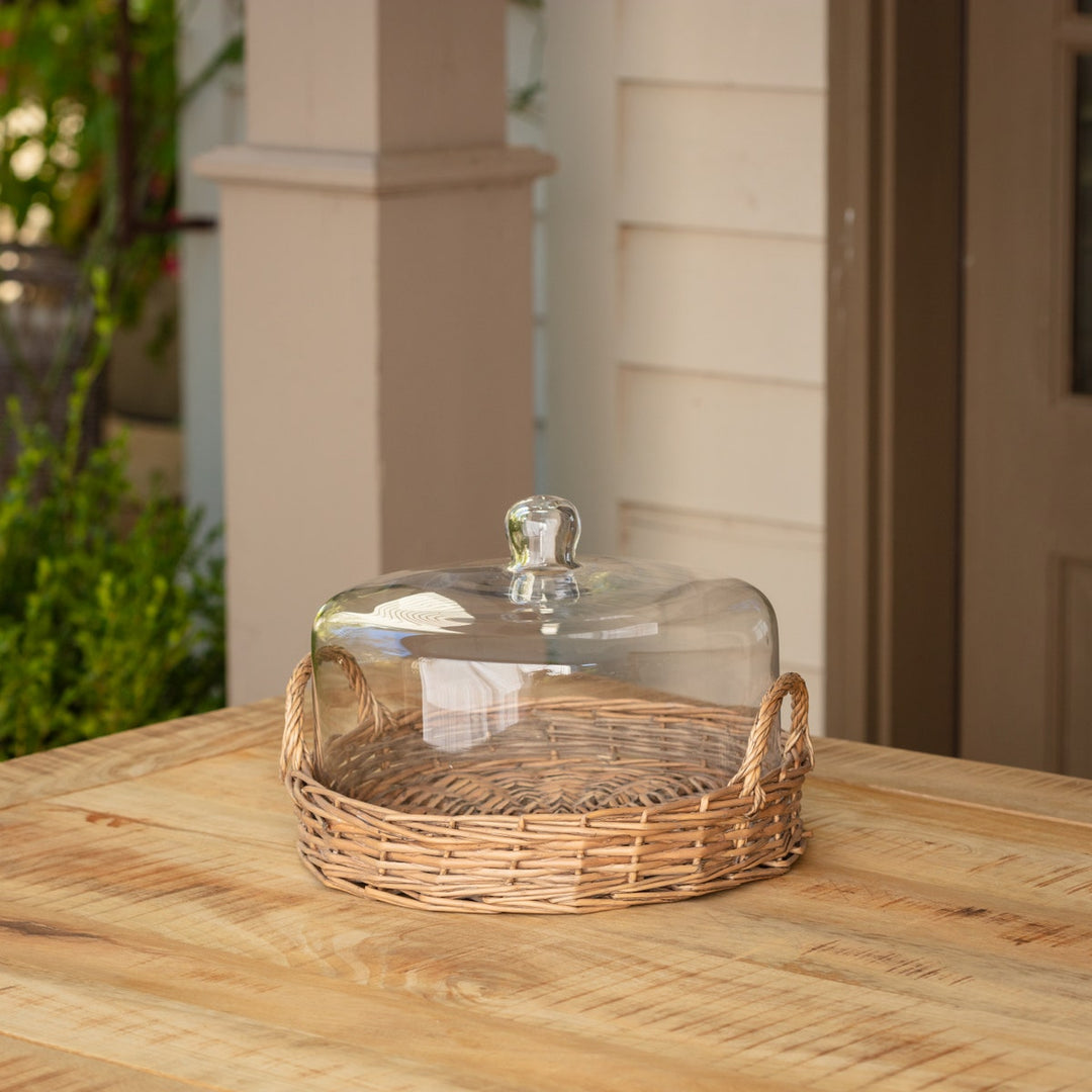 Large cloche covered basket with a clear glass dome and woven rattan base on a wooden table.