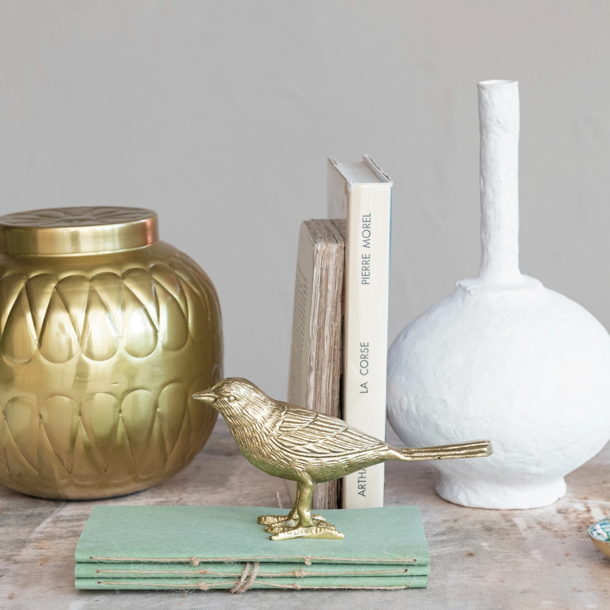Gold aluminum bird figurine standing on a stack of green books next to a vase and books.