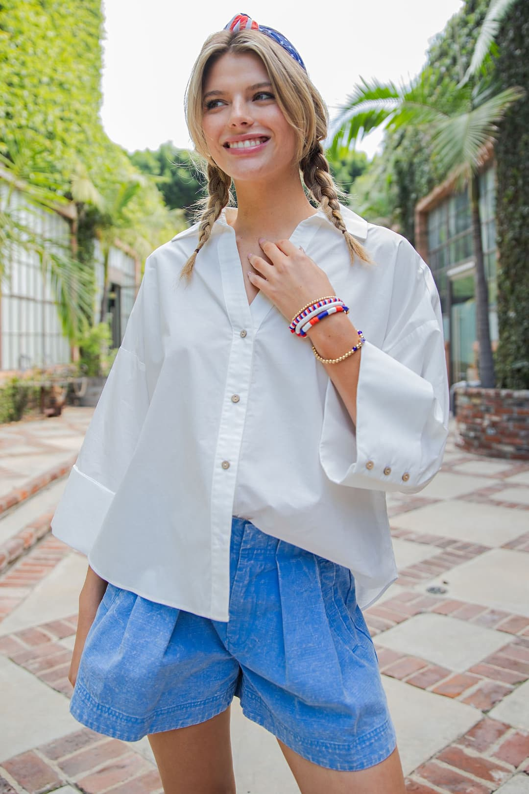 Woman wearing the white Blake Back Pleat Button Down Blouse with wide sleeves and blue shorts.