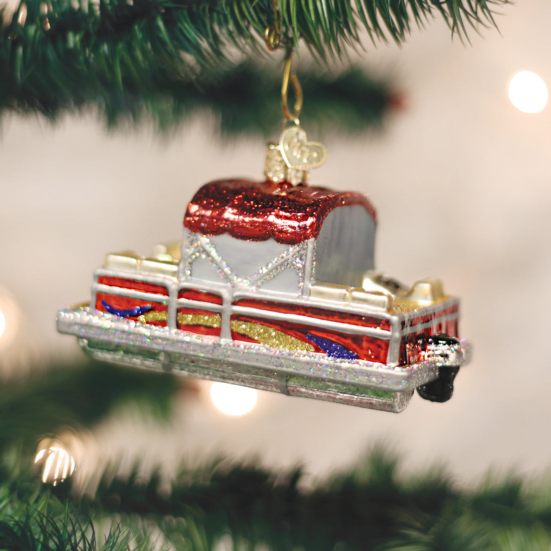 A glittery red and silver glass pontoon boat ornament hanging from a green Christmas tree branch.