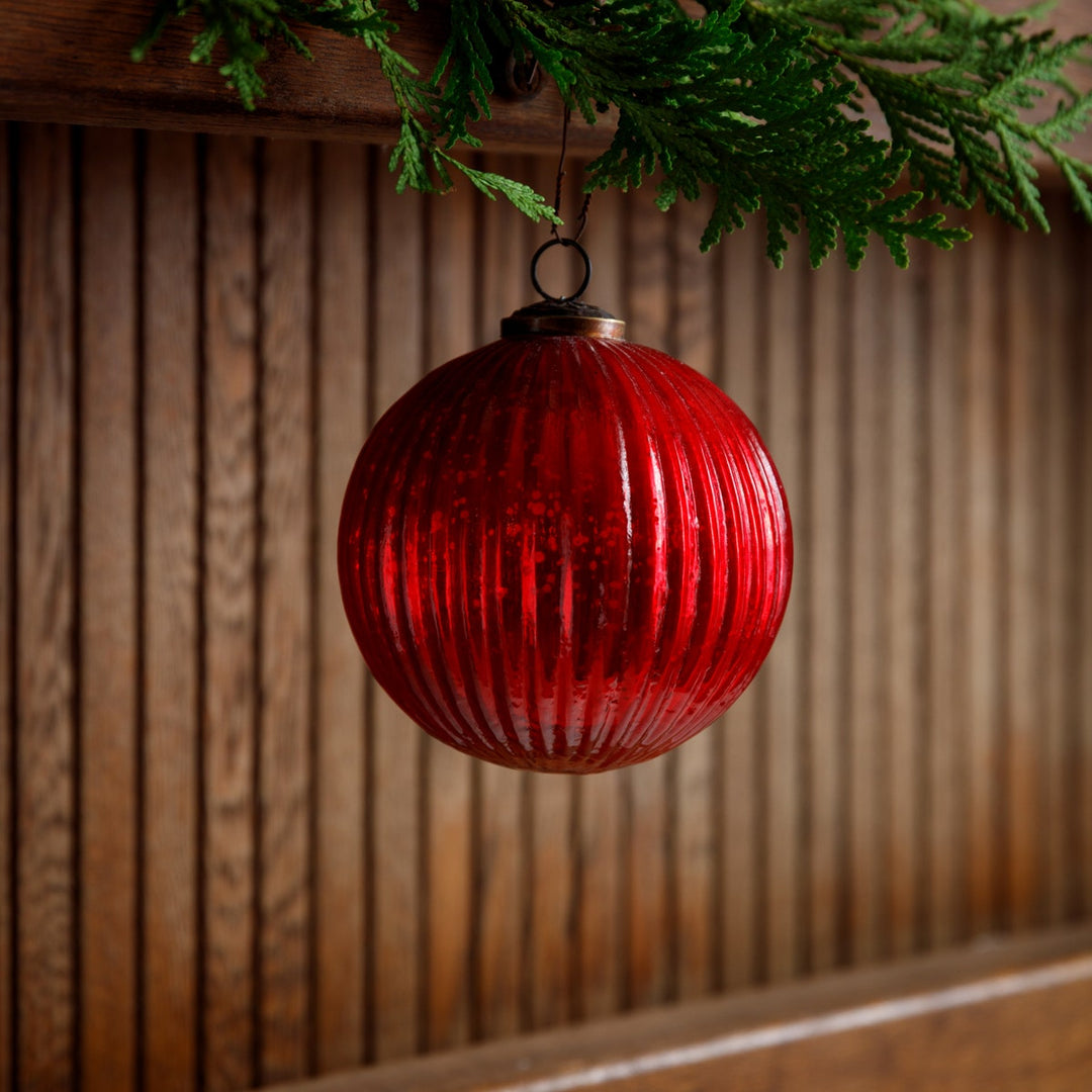 Red fluted glass ball ornament hanging from a green branch against a wooden background.