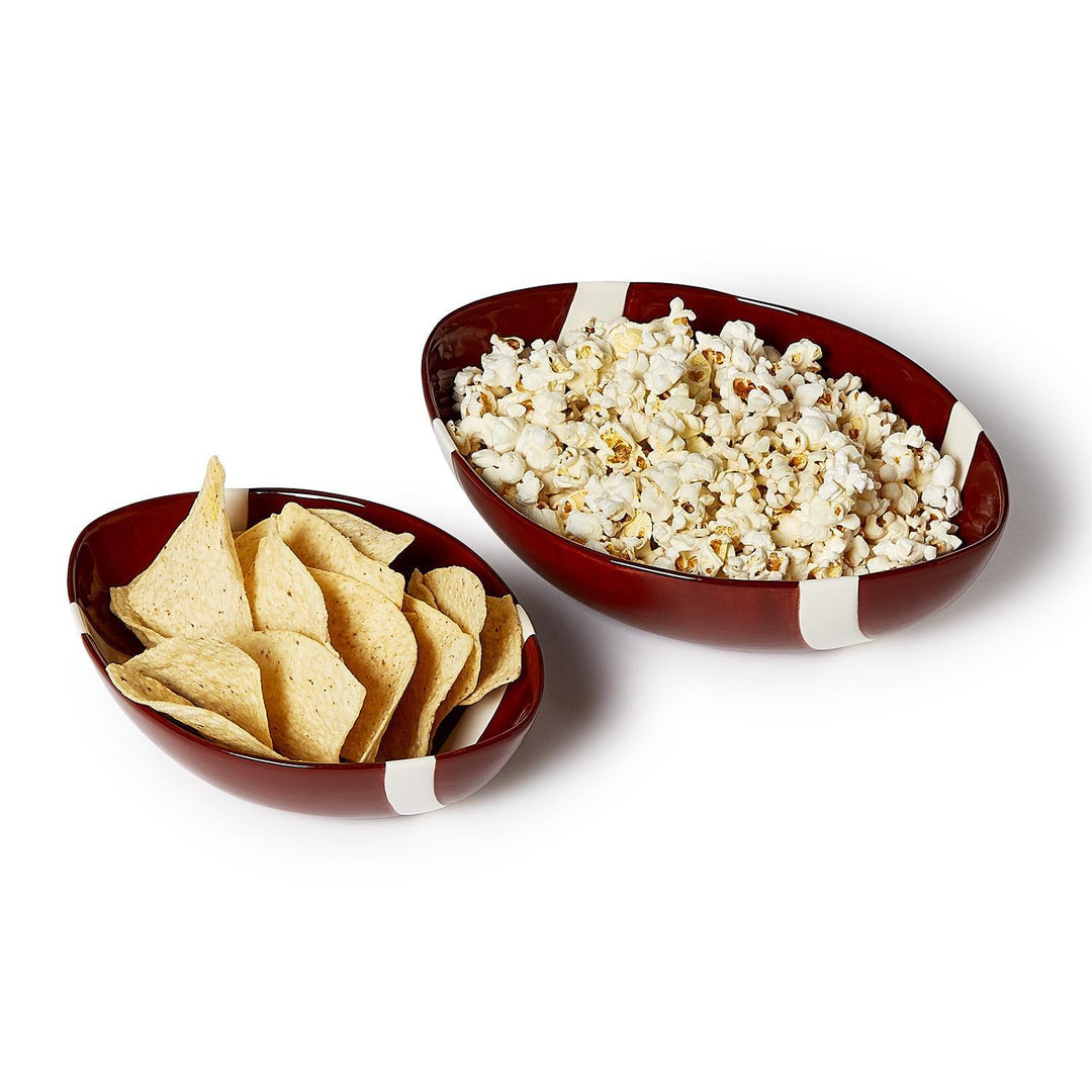 Two football-shaped ceramic bowls in different sizes, filled with popcorn and tortilla chips.