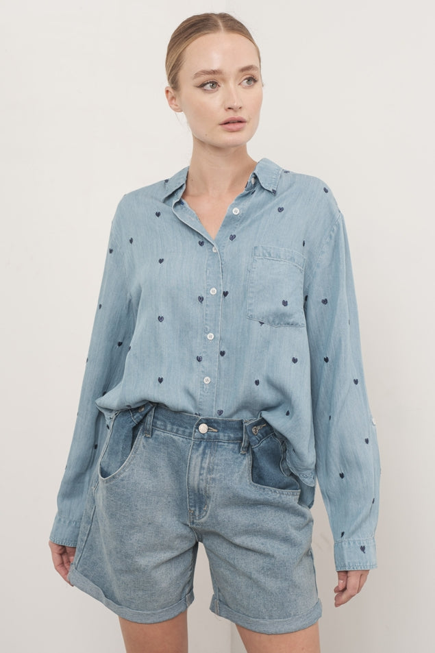 Woman wearing the Naomi Oversized Chambray Shirt with blue heart embroidery and denim shorts.