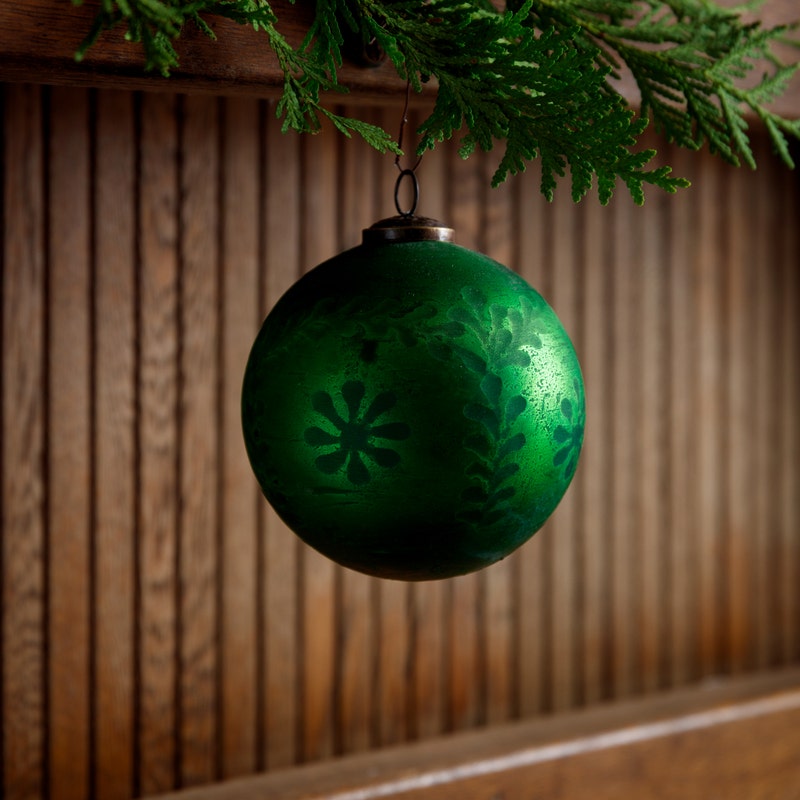 5-inch frosted green glass ball ornament with etched floral patterns hanging from a cedar branch.