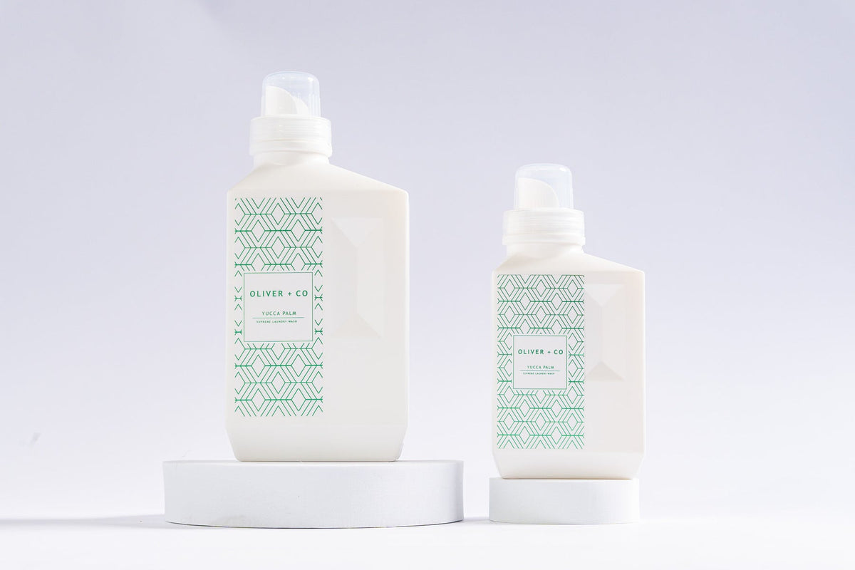 Two white bottles of Oliver + Co Supreme Laundry Wash in Yucca Palm on white circular pedestals.