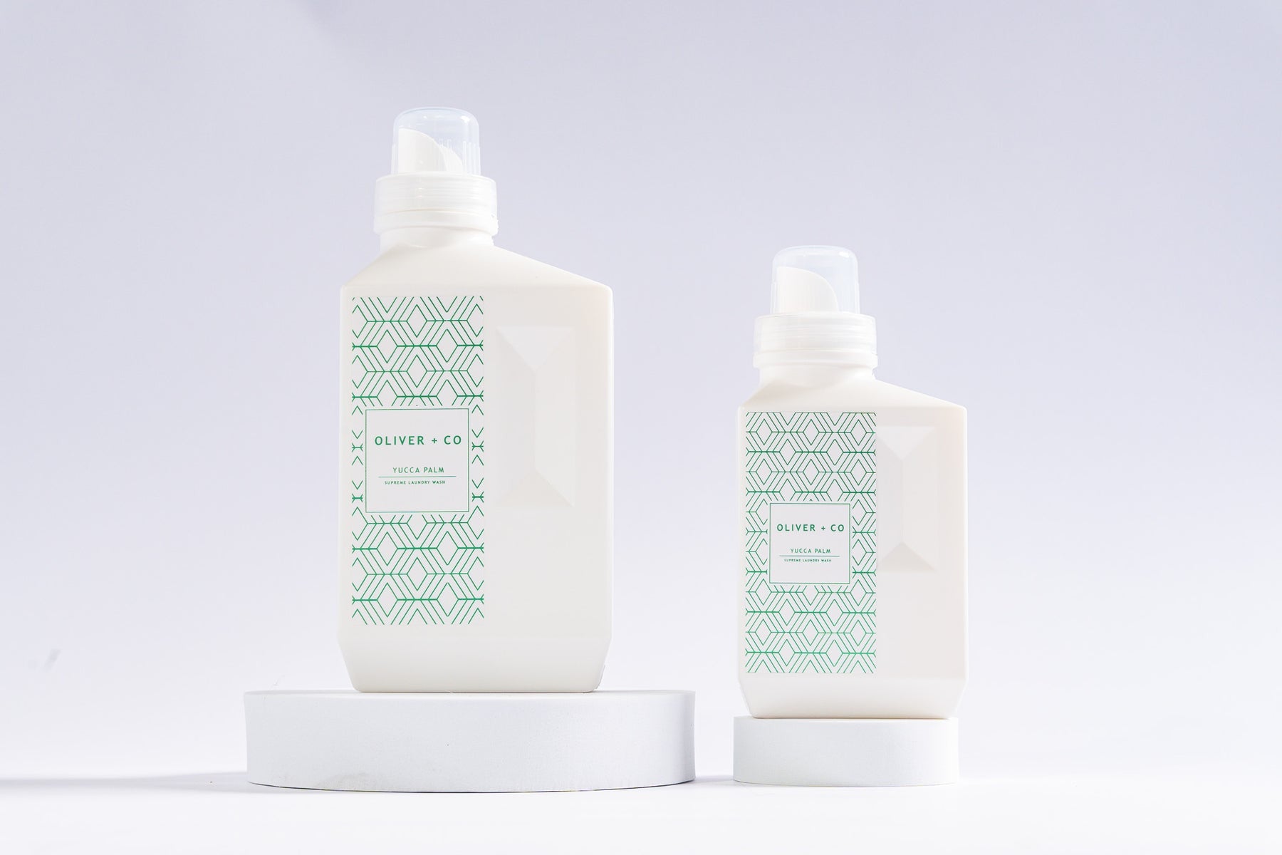 Two white bottles of Oliver + Co Supreme Laundry Wash in Yucca Palm on white circular pedestals.