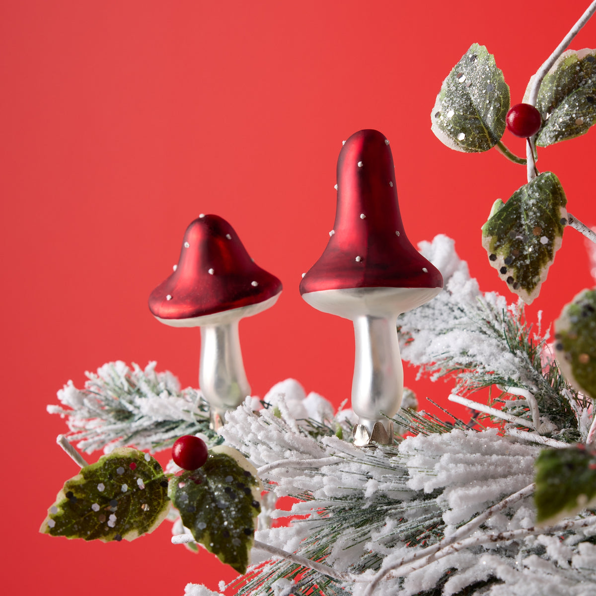 Two red and white glass mushroom clip ornaments on a snowy evergreen branch against a red background