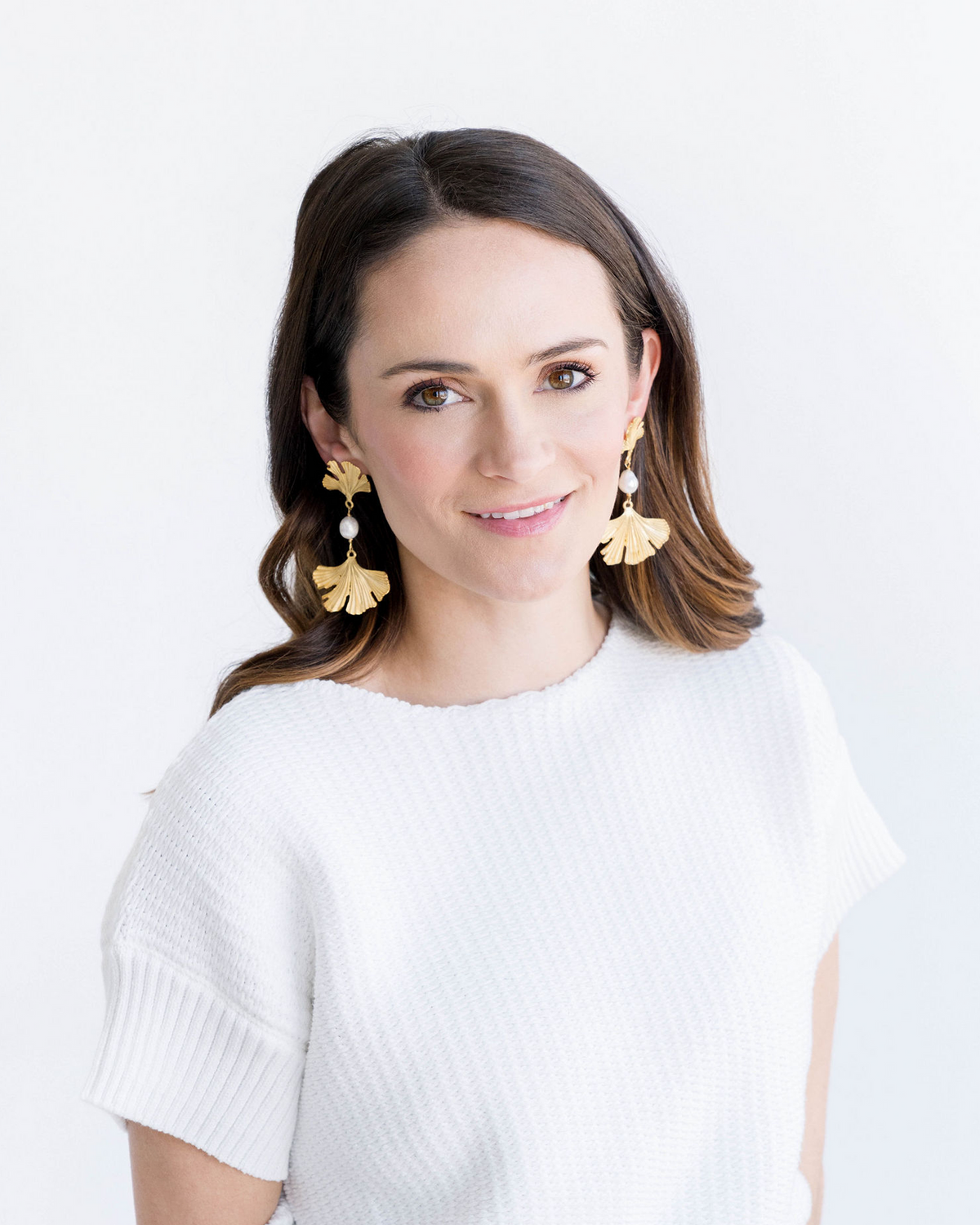 Woman wearing gold ginkgo leaf earrings with freshwater pearls and a white knit top.