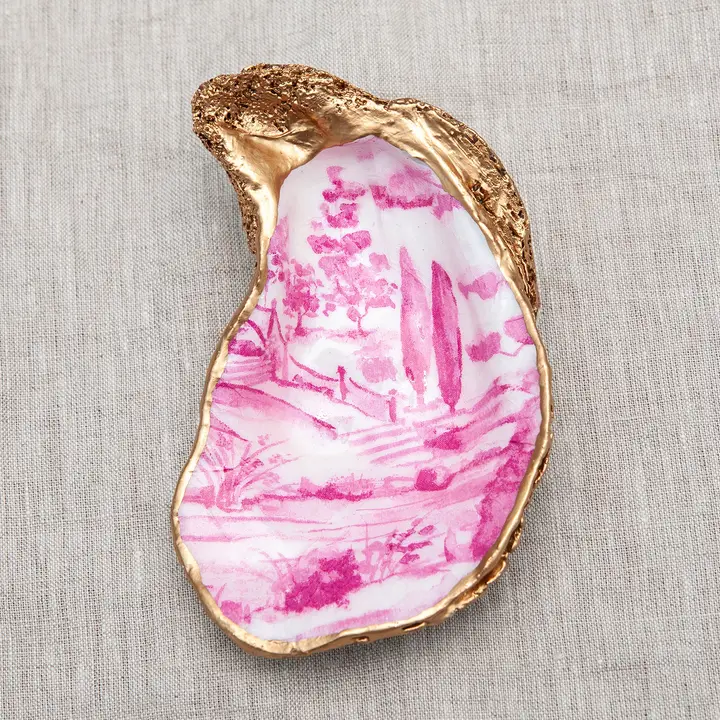 Decoupaged oyster shell with a pink landscape scene and metallic gold trim on a linen background.