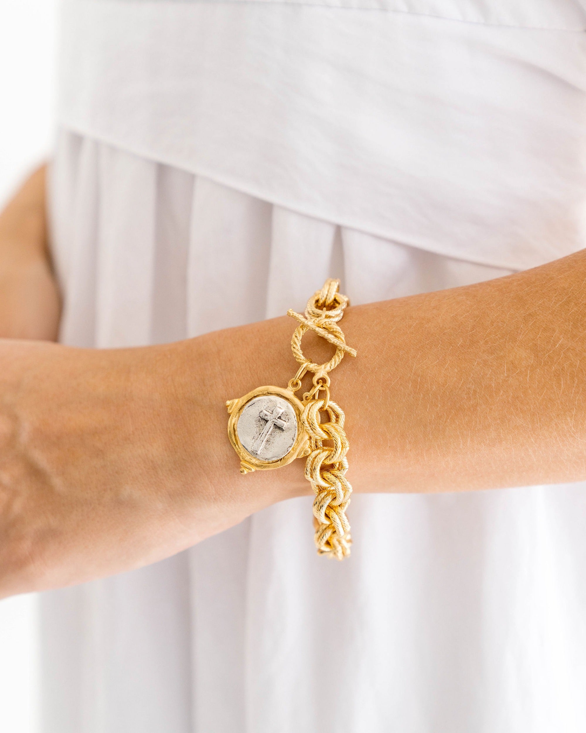 A person wearing a gold chunky chain bracelet with a silver cross coin charm and toggle closure.