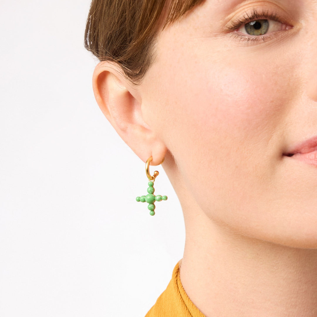 Close-up of a person wearing a gold hoop earring with a green beaded cross charm.