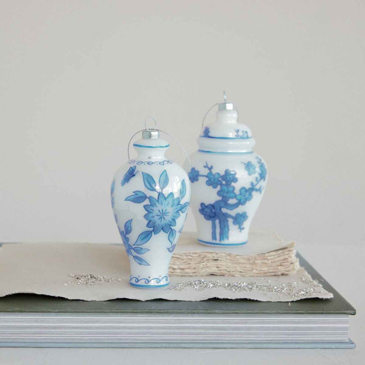 Two white glass ginger jar ornaments with hand-painted blue floral motifs on a stack of books.