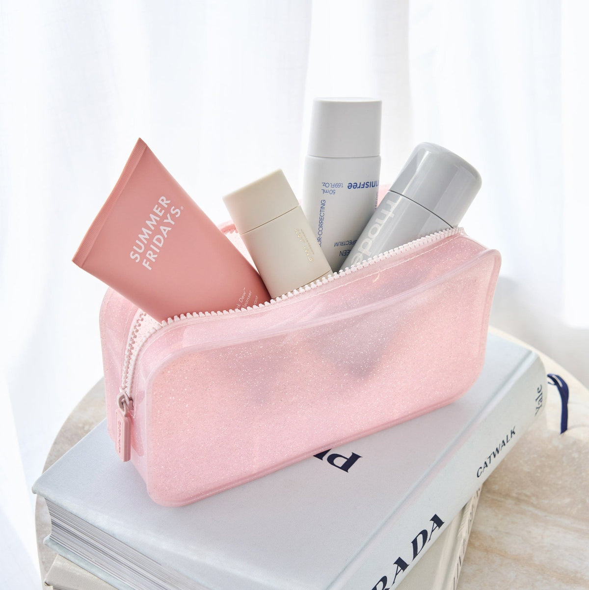 Jelly Makeup Bag in Ballet Pink Twinkle filled with skincare products on a stack of books.