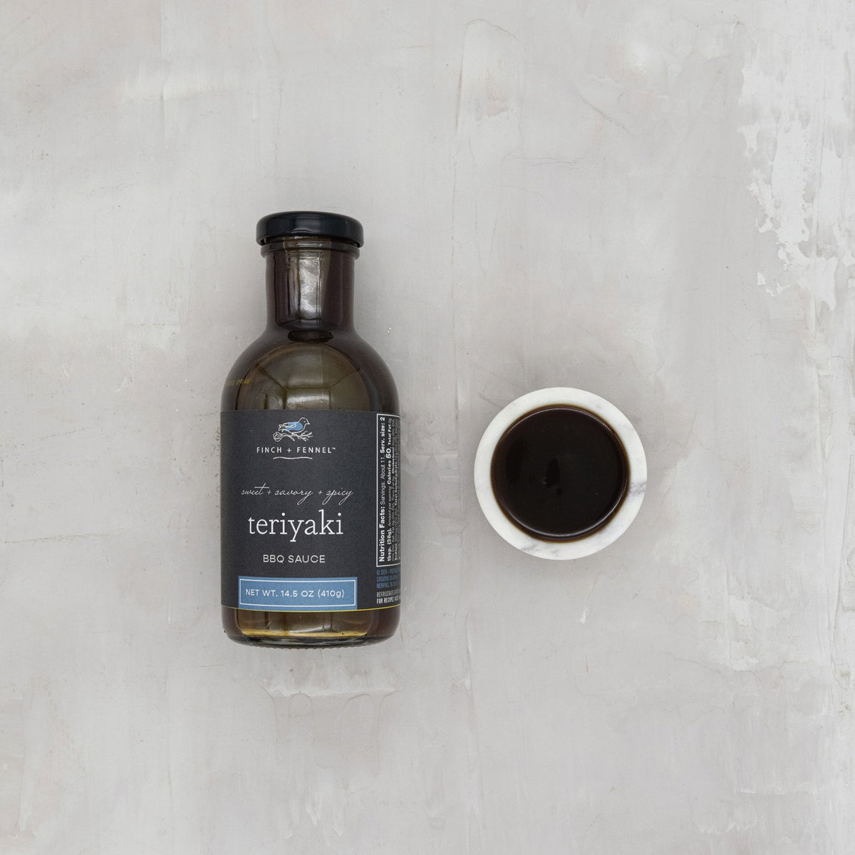 A bottle of Finch + Fennel Teriyaki BBQ Sauce next to a small white bowl filled with the sauce.