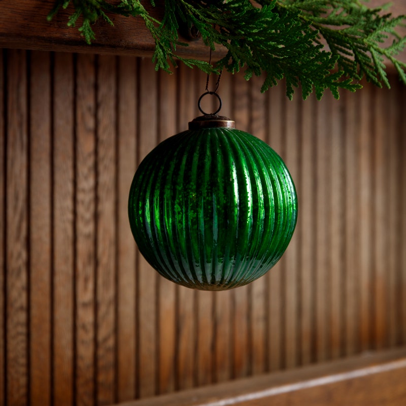 Green fluted glass ball ornament hanging from a cedar branch against a wood panel background.