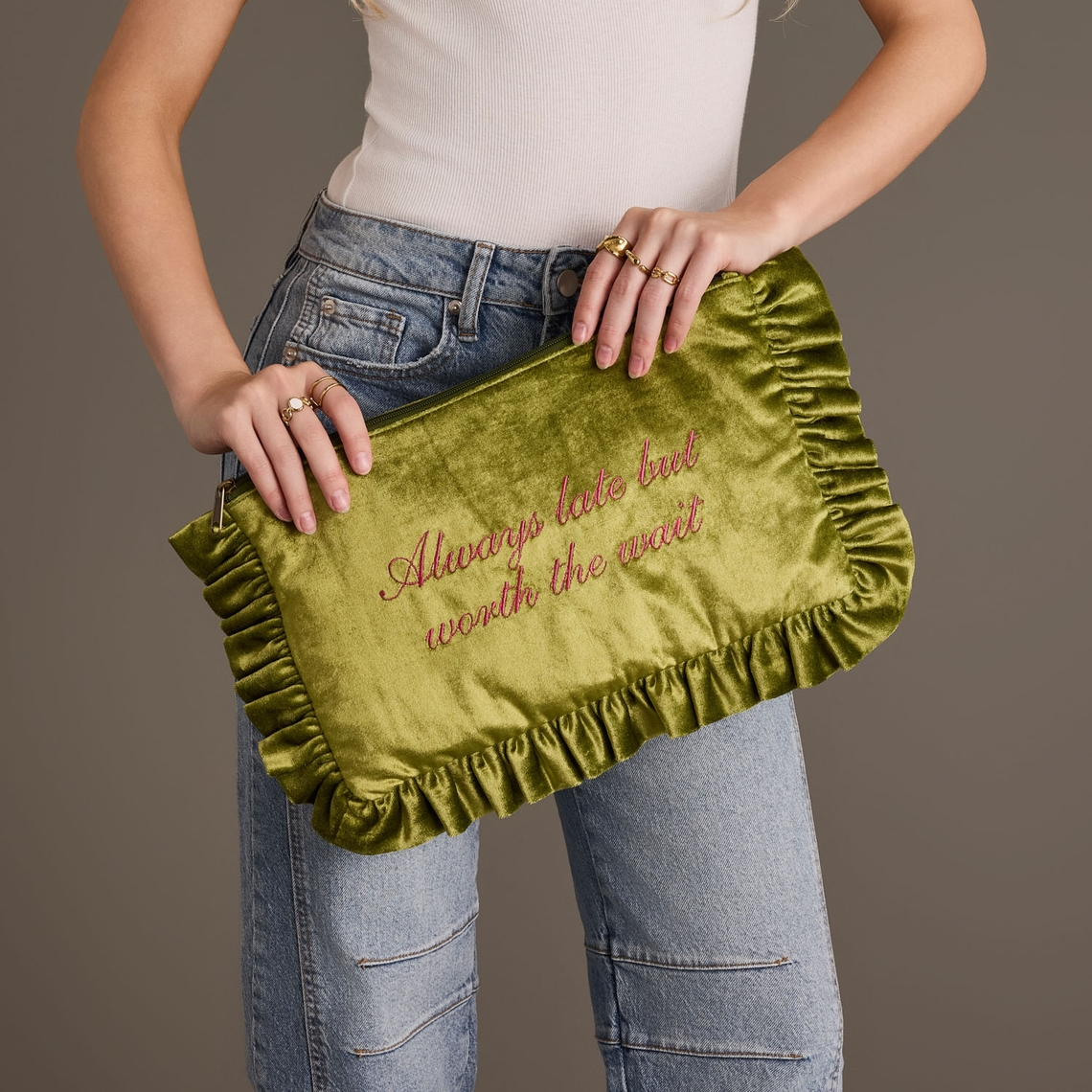 Green velvet ruffle travel pouch with pink embroidery reading Always late but worth the wait.