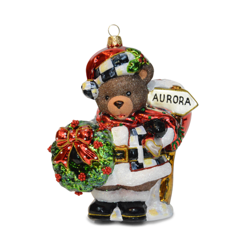 Aurora Teddy Bear glass ornament wearing a checkered hat and coat, holding a wreath and sign.