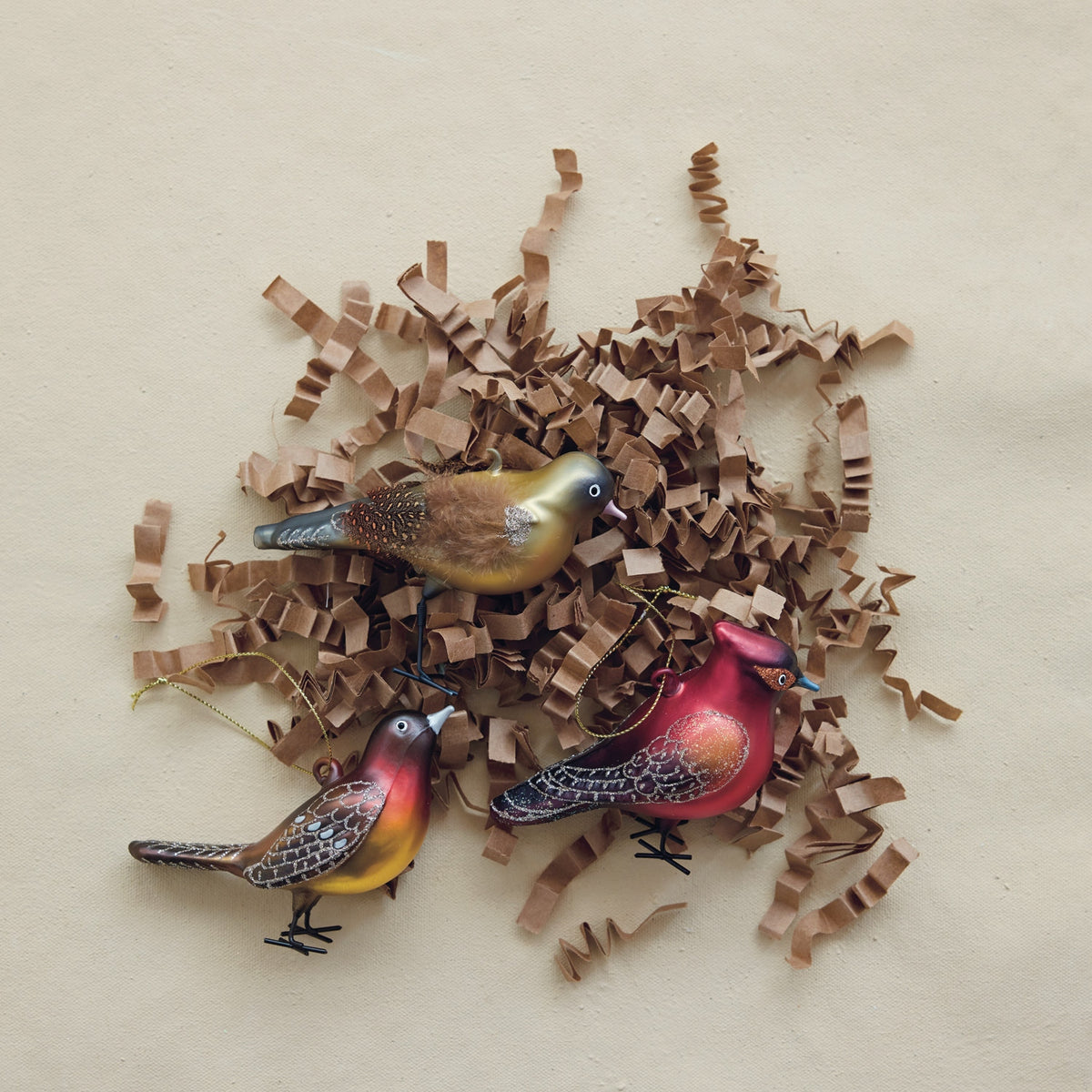 Three hand-painted glass bird ornaments with glitter wings on brown shredded paper.