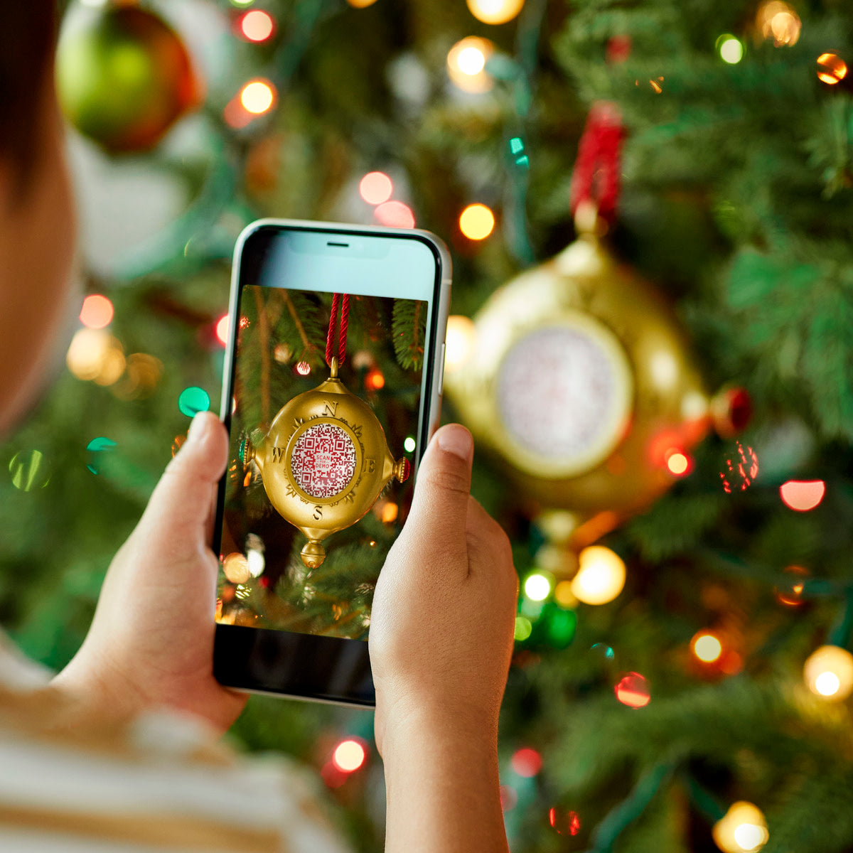 A child uses a smartphone to scan a QR code on a gold compass-shaped Santa's Kindness Ornament.