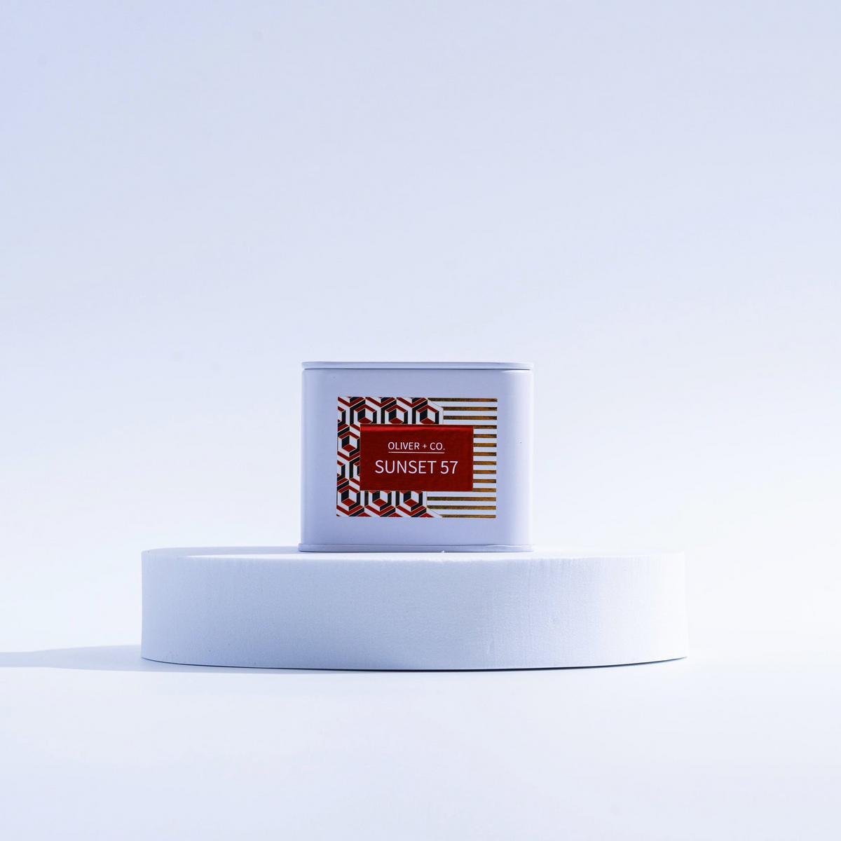Oliver + Co. Sunset 57 Travel Companion Candle in a white tin on a circular white pedestal.